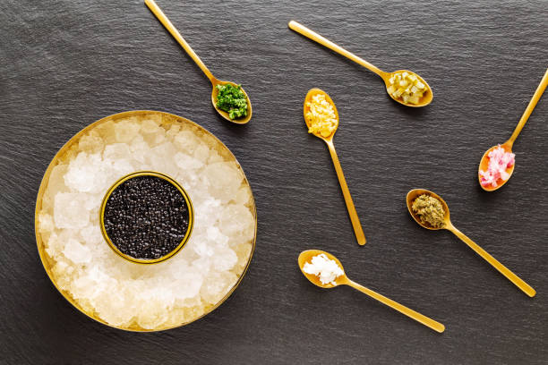 High quality real black sturgeon caviar in golden box on crushed ice aside golden spoons with pickles and herbs on a slate background. Flat Lay. Top view. stock photo