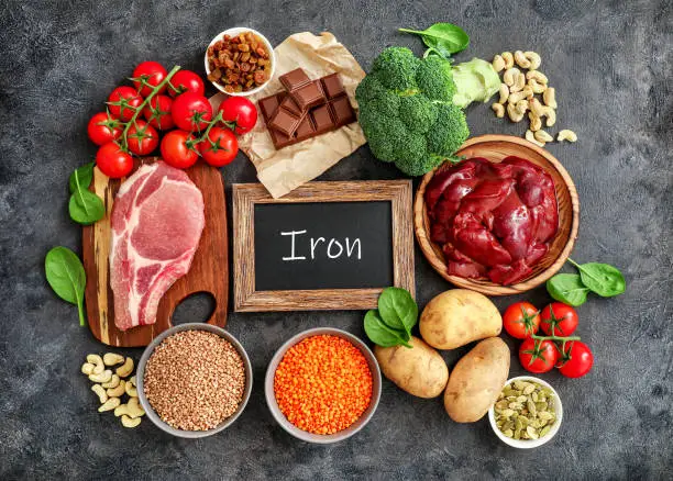 Assortment of high in Iron sources on dark background: liver, beef, raisins, keshew, buckwheat, spinach, tomatoes, potatoes, dark chocolate, pumpkin seeds, lentil, broccoli. Top view.