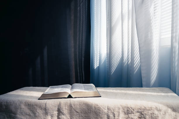 istock Open Holy Bible  on a white table, black wall and window  background. Stay at home. Religion concept. 1256640324