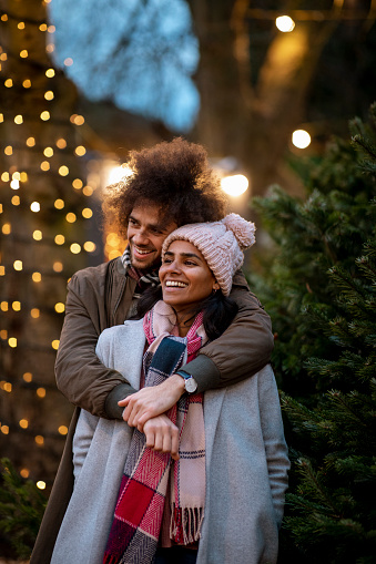 A close-up of a multi-ethnic couple standing in a Christmas tree farm together in Newcastle-Upon-Tyne. The man is standing behind his wife with his arms around her, they are looking away from the camera and smiling.