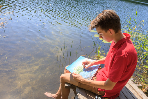 Teenager boy sitting on a wooden pier by forest lake drawing with pastel crayons - plein air scetching concept
