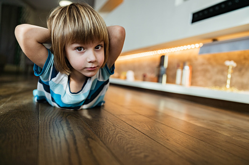 Cute little boy doing back exercises with hands behind his head on parquet floor at home and looking at camera. Copy space.
