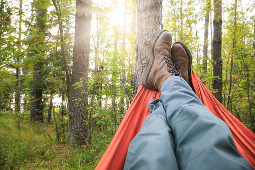 Person wearing trekking pants and hiking boots relaxing laying in camping hammock overlooking summer forest at sunset