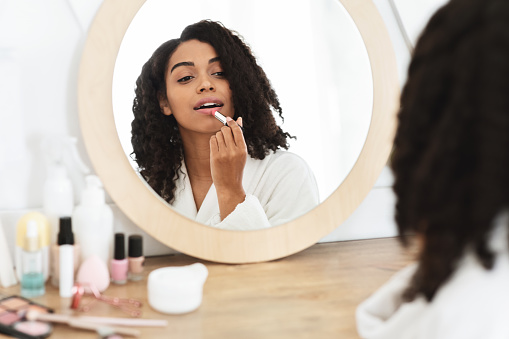 Beauty Portrait Of Attractive Black Girl Applying Pink Nude Lipstick In Front Of Mirror