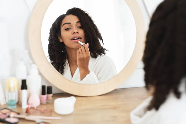 beauty portrait of black girl applying pink nude lipstick in front of mirror - mirror women bathroom make up imagens e fotografias de stock