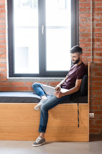 istock handsome businessman using laptop while sitting on window bench in modern office 1256678290