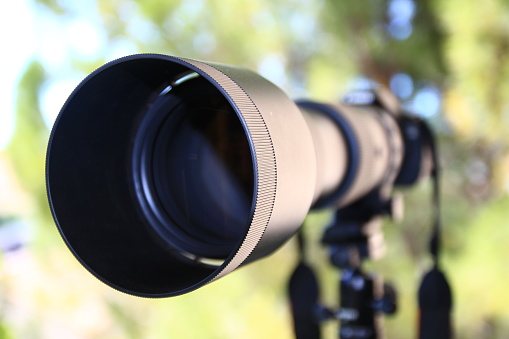 A three quarter view of an imposing telephoto lens and its sun visor, fixed in front of a camera. The strap and the tripod are seen in the background.