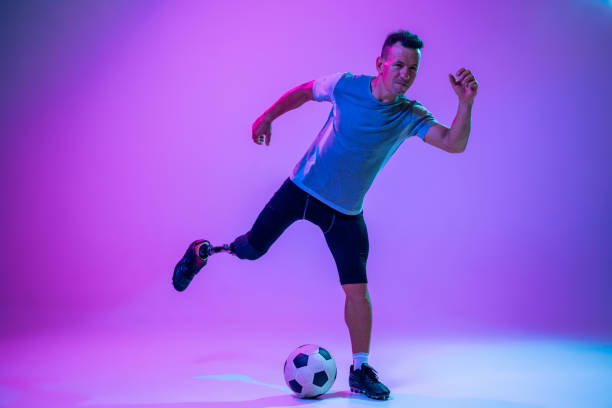 istock Athlete with disabilities or amputee isolated on gradient studio background. Professional male football player with leg prosthesis training and practicing in studio. 1256690749