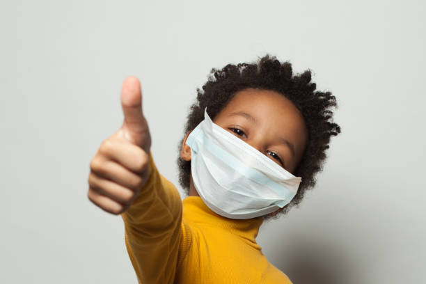 enfant noir afro-américain heureux dans le masque protecteur médical de visage affichant le pouce vers le haut sur le blanc - safety protective workwear healthcare and medicine healthy lifestyle photos et images de collection