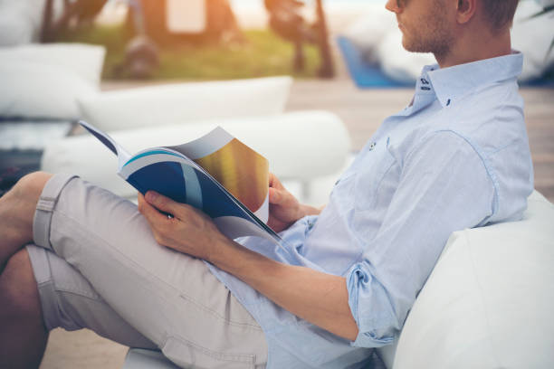 joven caucásico leyendo el libro de la revista sobre el lector de sofás sosteniendo una página de papel abierta del libro y leyendo mientras se sienta al aire libre en el sofá fuera de la biblioteca y el libro de lectura. estilo de vida para el hombre. - e reader cheerful indoors lifestyles fotografías e imágenes de stock