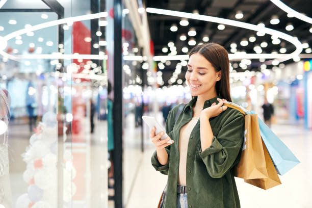 Young cheerful brunette shopper with paperbags scrolling in smartphone Young cheerful brunette shopper with paperbags scrolling in smartphone or looking through messages or adverts while shopping in the mall young women shopping stock pictures, royalty-free photos & images