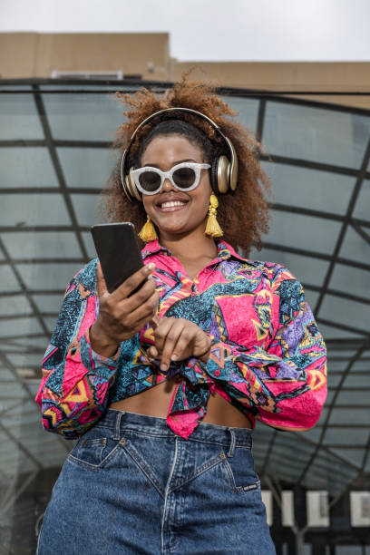 stylish black woman in headphones surfing internet on smartphone outdoors - surfing wireless vertical outdoors lifestyles imagens e fotografias de stock