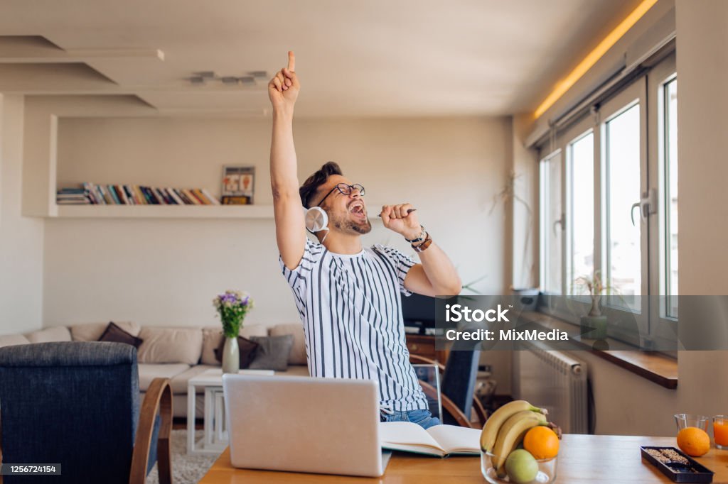 Excited young man making his own crazy party at home Excited young man dancing and singing in front of his laptop at home. Humor Stock Photo