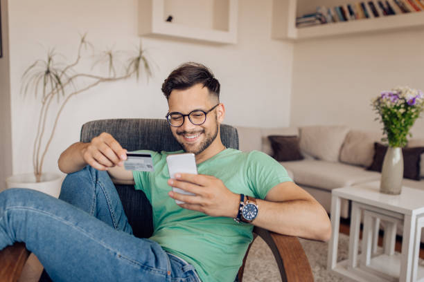Happy young man shopping online at home Young man relaxing and shopping online at home. credit card purchase stock pictures, royalty-free photos & images
