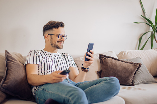 Young man sitting on the sofa, using a smartphone and and watching TV at home.