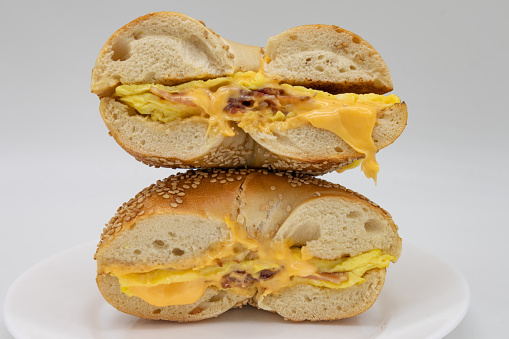 A sesame seed bagel cut in half and stacked while filled with bacon eggs and cheese on a white plate with a white background