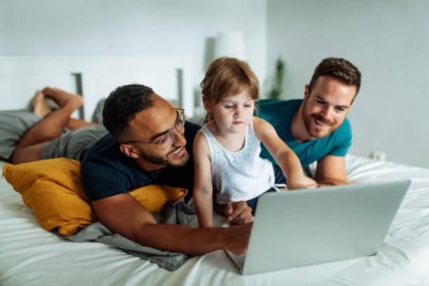 LGBT couple watching videos on laptop with adopted daughter Gay couple playing with their adopted daughter girl and enjoying the parenthood gay daddy stock pictures, royalty-free photos & images