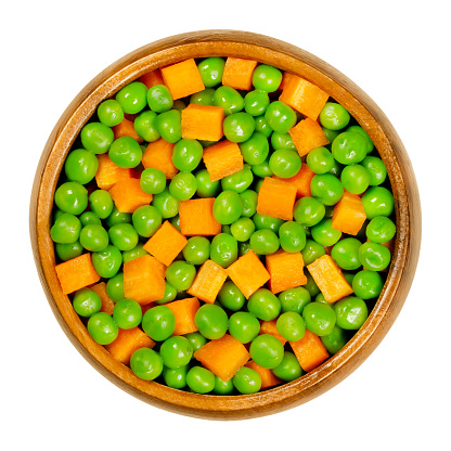 Green peas and carrot cubes in wooden bowl. Mixed vegetables. Seeds of pod fruit Pisum sativum and orange colored cubes of carrots, Daucus carota, a root vegetable. Closeup from above macro food photo