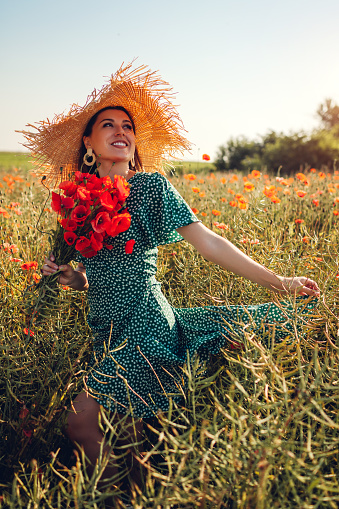 Portrait of woman gathered bouquet of poppies flowers walking in summer field. Stylish girl in straw hat admires view. Vacation