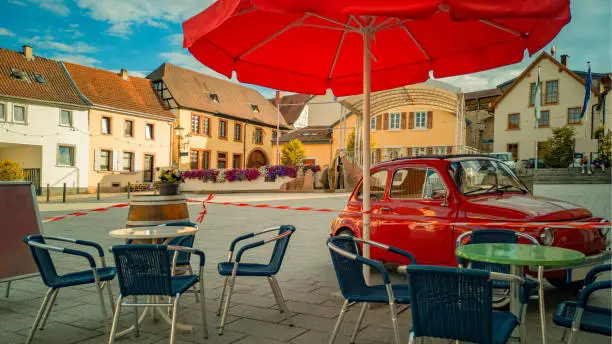 Terrace of street cafe with empty tables,wicker chairs and parasole,fenced off by red-and-white barrier tape,with red mini-car parked nearby,on German old square in center,Europe,on bright Sunny day.