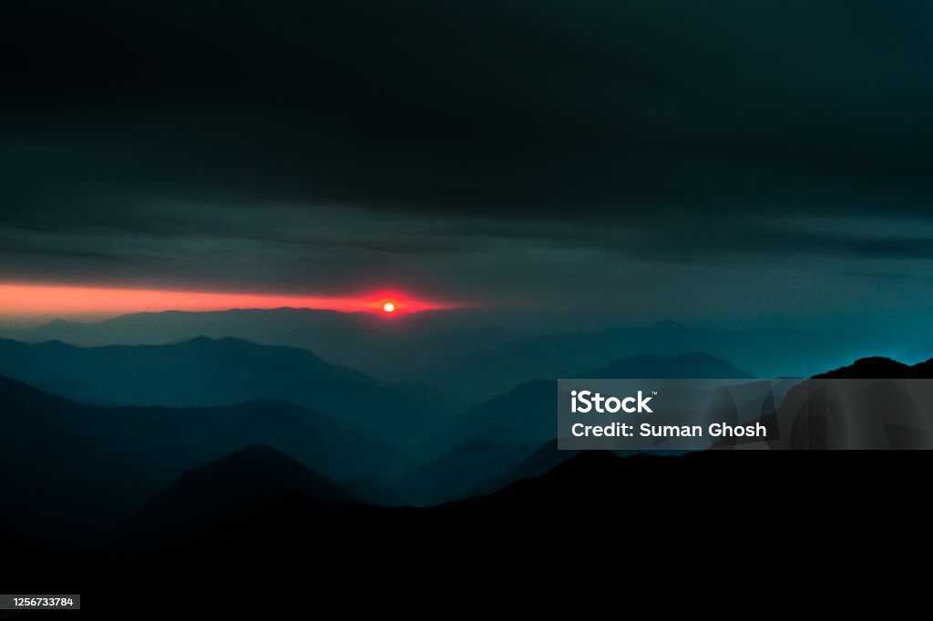 A landscape of the layers of hills with the setting sun in the background and dark cloudy sky above Atmospheric Mood Stock Photo
