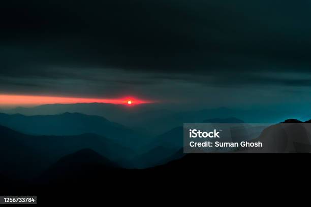 A Landscape Of The Layers Of Hills With The Setting Sun In The Background And Dark Cloudy Sky Above Stock Photo - Download Image Now