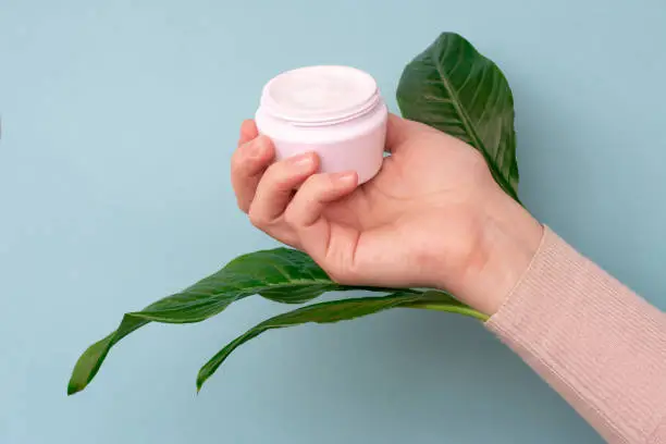 Young girls hand is holding a jar with white soft cream on pale blue background with large green leaves.Concept of eco cosmetic and beauty procedure.