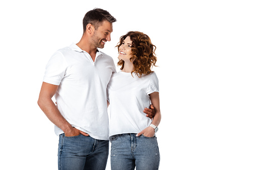 happy man and woman standing with hands in pockets while looking at each other isolated on white