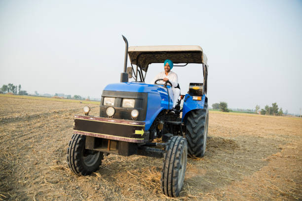A weathered tractor sits in a rural field under a clear blue sky. The tractor is covered in rust, and its tires are worn down. The surrounding field is covered in dirt and grass, and there are other tractors in the distance.