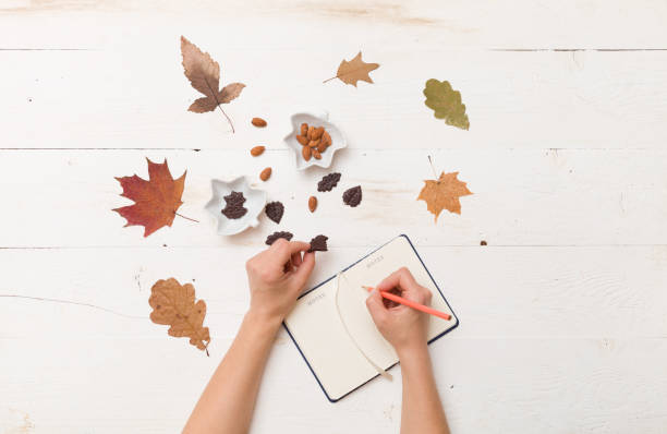 istock Top view on woman's hands writing in notebook. Autumn background: leaves, chocolate, nuts on wooden white board. 1256743800