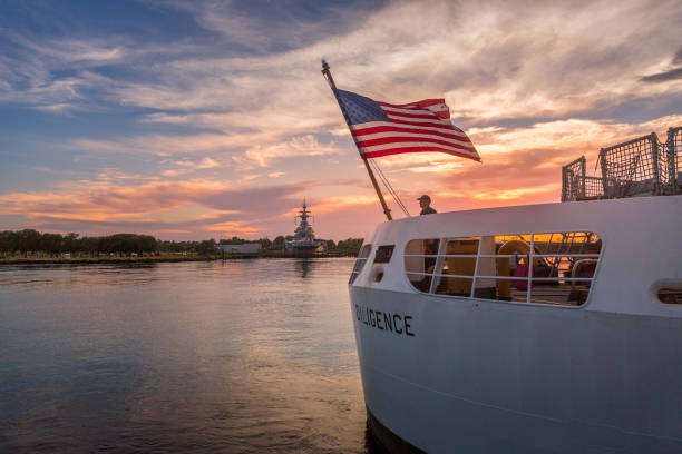The sun sets behind the American flag and a sailor aboard the USCGC Diligence stock photo