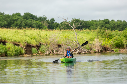 Front view of a young girl kayaking on a small lake on a summer day. The girl is wearing a straw hat and blue life jacket. She is in a green and yellow kayak with a black paddle.