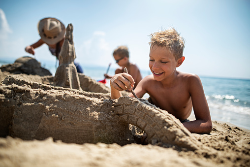 Kids having fun building sandcastles on beach. The boy is carving a sand bridge to his sand castle.\nNikon D810