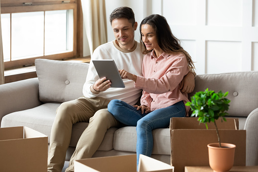 Happy young couple using computer tablet together, looking at mobile device screen, sitting on couch in living room with cardboard boxes with belongings, choosing moving service, relocation concept