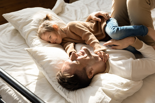 Happy father and little daughter relaxing in cozy bed together, smiling dad having fun with cute preschool girl, resting, taking nap in bedroom, family spending weekend together, enjoying free time