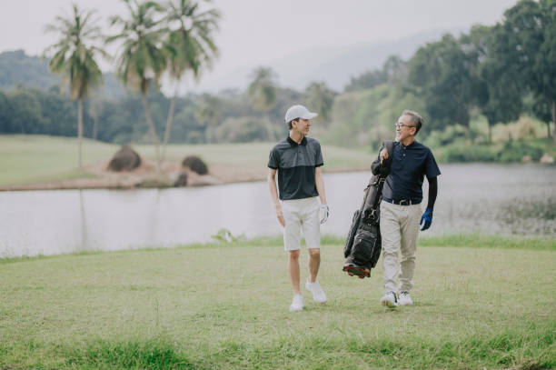 ein asiatischer chinese mit seinem teenager-sohn zu fuß in richtung golfplatz für das spiel tragen golftasche blick auf die aussicht - retirement golfer happiness relaxation stock-fotos und bilder