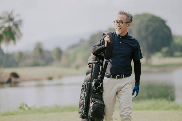 a matured chinese man golfer carrying a golf bag looking away in the golf course side view of a matured man golfer carrying a golf bag looking away in the golf course rich man stock pictures, royalty-free photos & images