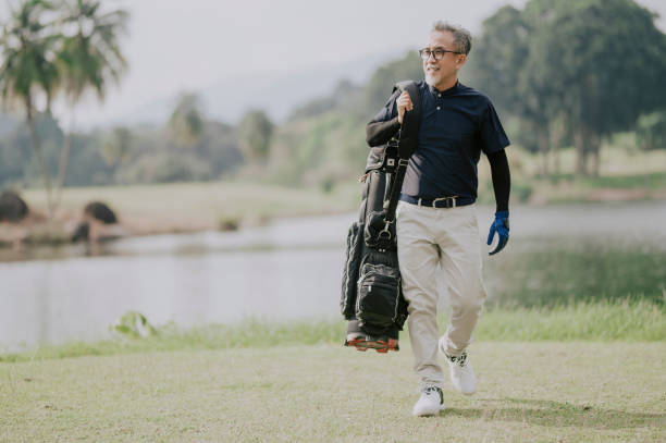 un golfista chino maduro que lleva una bolsa de golf mirando hacia otro lado en el campo de golf - retirement golfer happiness relaxation fotografías e imágenes de stock