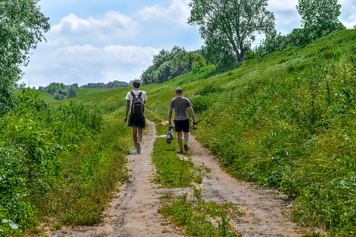 Two fishermen are walking along a meadow dirt road, view from the back. Male hikers with backpack and fishing rods among green nature on a sunny spring-summer day