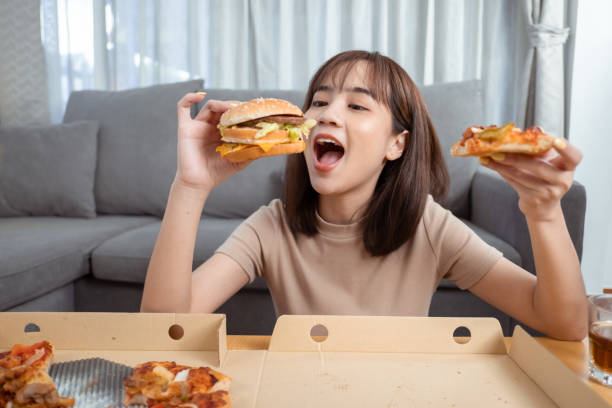 close up asian woman takeaway eating junk food hamburger and pizza close-up shot in living room at home,  fast food delivery service safe and stop coronavirus spread by social distancing concept. - burger hamburger large food imagens e fotografias de stock