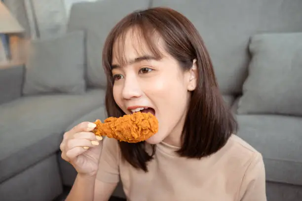Photo of Young asian woman takeaway eating junk food chicken nuggets wing, Female having fun enjoying fast food delivery service safe and stop coronavirus spread by social distancing concept.