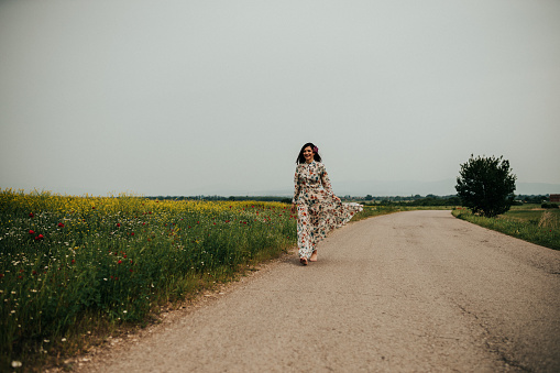 Young woman wearing a floral dress and walking on the road next to an agricultural field on a sunny summer day