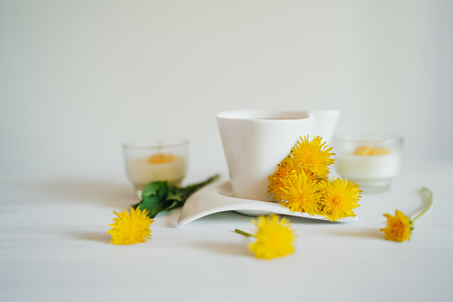 Cup of healthy dandelion tea with fresh dandelion flowers and yellow candles on white background. Herbal medicine and aroma therapy concept