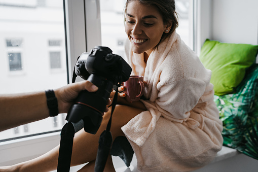 Beautiful young woman at home, sitting on window sill and looking camera