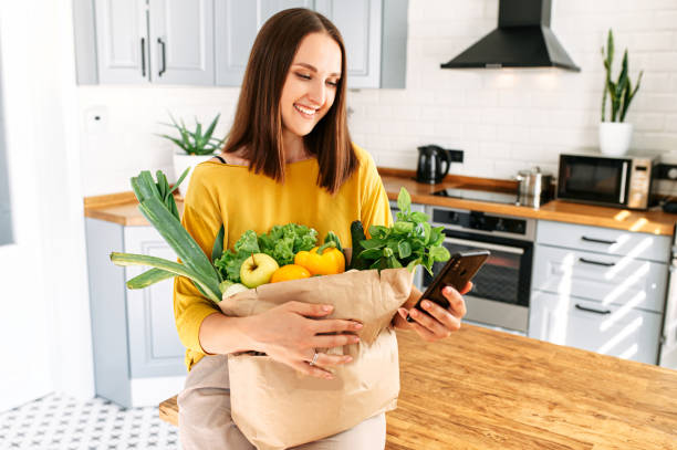 女性は、携帯電話と野菜でいっぱいのバッグを保持します - paper bag groceries food vegetable ストックフォトと画像