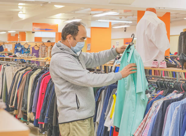 istock Mature man in mask choosing clothes at Second hand store. 1256843028