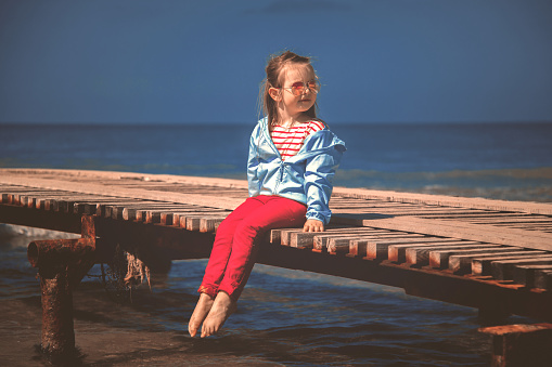 Charming little fashionista is resting by the lake, wearing a blue jacket and a striped t-shirt and trendy red jeans.