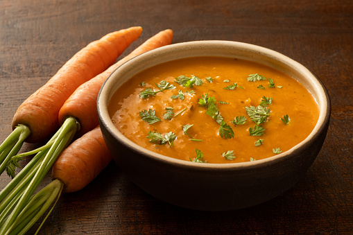 Carrot and coriander soup with freshly chopped coriander in a brown rustic bowl next to three whole carrots.
