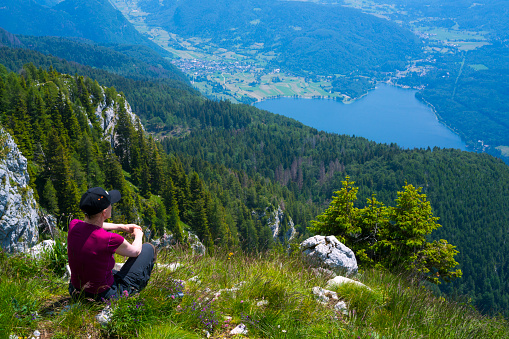 Senior woman sitting on the edge of hill high up and enjoying the view of Lake Bohinj panorama   and the mountains around, Slovenia.