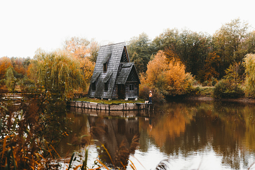 Young woman in hat and her small dog enjoying the beautiful and colorful autumn day staying at the pier near the wonderful old abandoned fishing house at the reflection lake